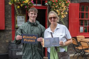 This is a mother and son holding up a placard and posing for the camera. The placards have written on them a charity message which says "1 in 3 children in crises-affected countries are out of school" and "UnitetoProtect". They are standing in front of a door with flowers hanging from it, near Borough Market in London.